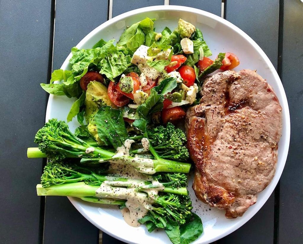 A white plate with a piece of meat with broccoli, tomatoes, cheese, avocado and lettuce