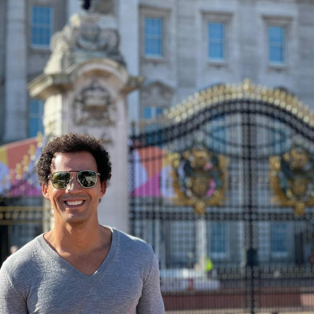 Pericles Rosa wearing sunglasses and a grey t-shirt posing in front of the Buckingham Palace main gate, London