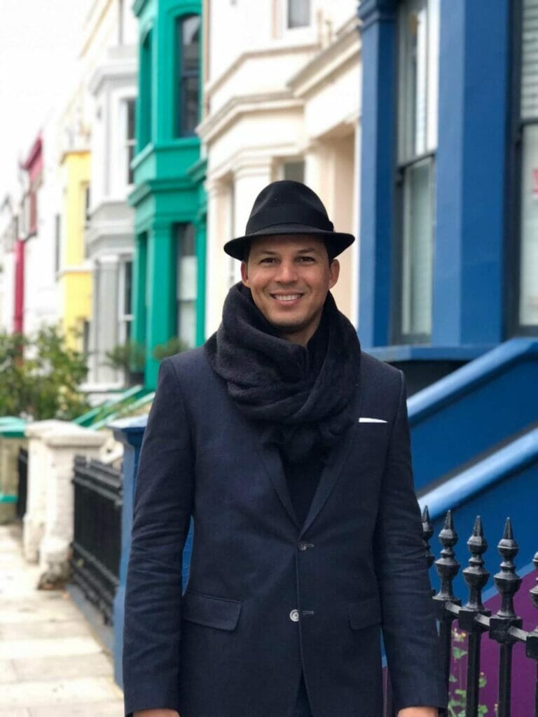 A man wearing a black hat, black scarf and blue blazer at Lancaster Road in Notting Hill, London, and its colourful houses in the background