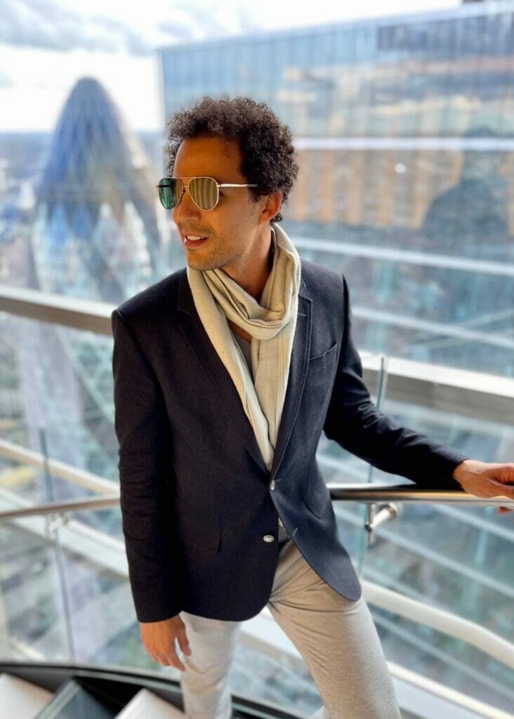 Pericles Rosa wearing sunglasses, a scarf, blue blazer and trousers at SushiSamba Restaurant, London, with some building, including The Gerkin, in the background