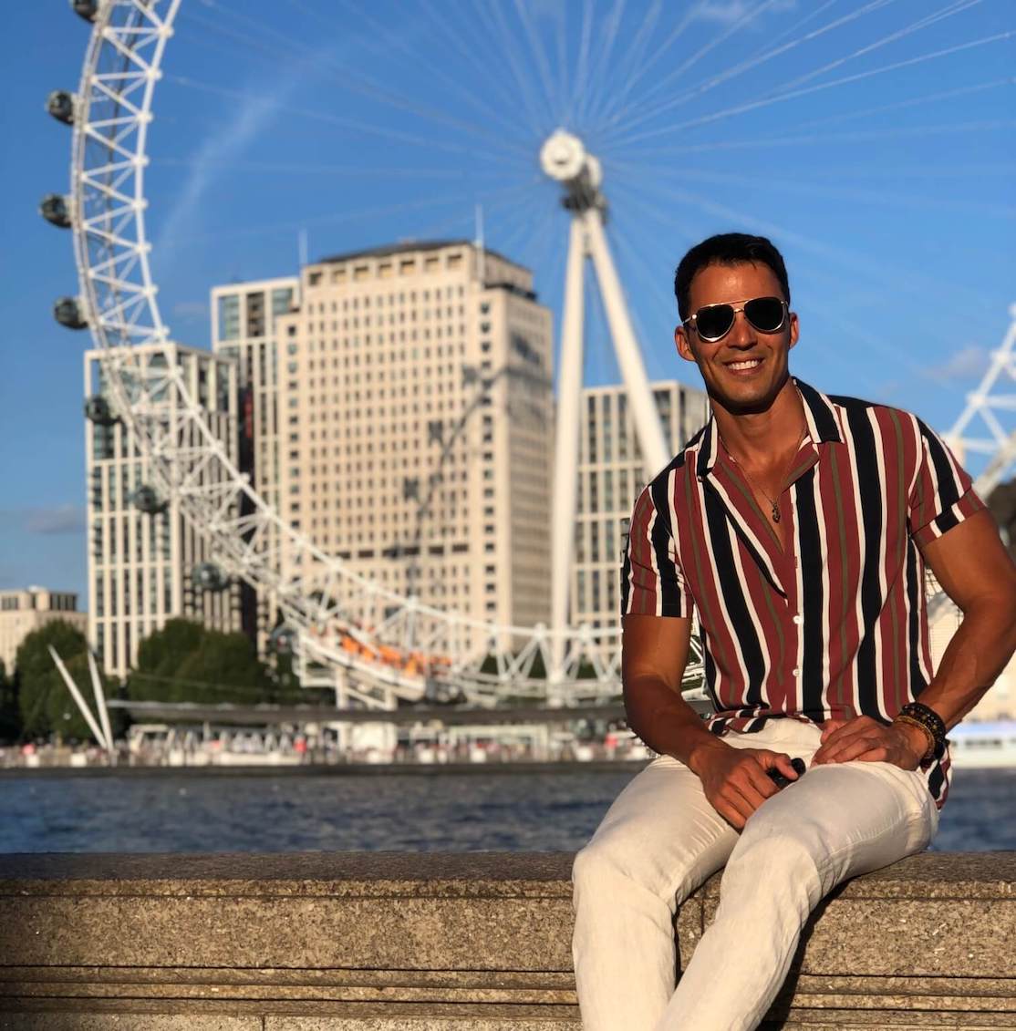 Pericles Rosa wearing sunglasses and stripe shirt posing to take a picture along the River Thames with the London Eye in the background