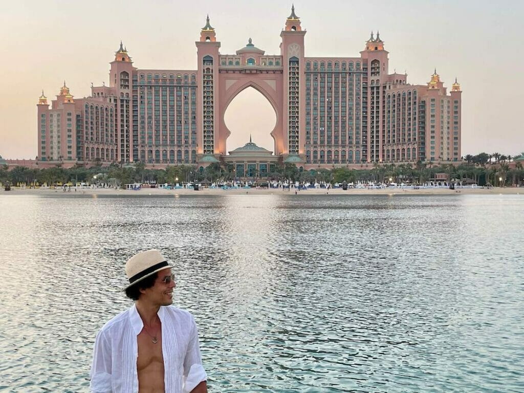 Péricles Rosa usando óculos escuros, um chapéu bege e uma camisa branca na praia do The Pointe em Dubai com o Hotel Atlantis ao fundo