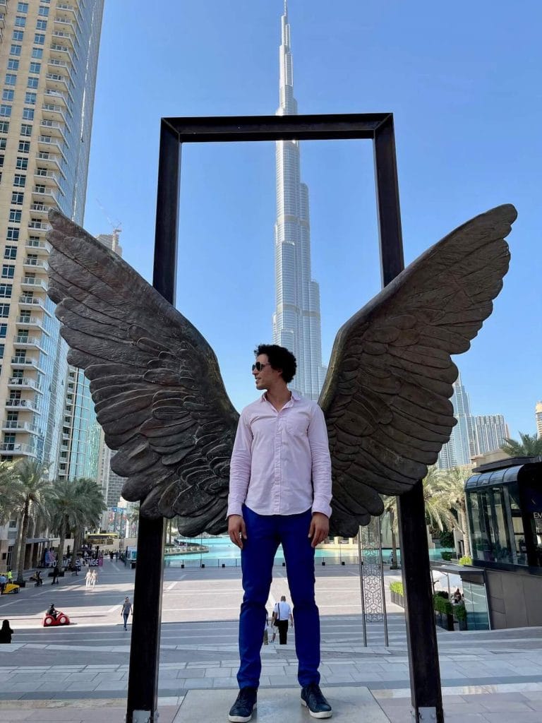 Pericles Rosa vistiendo una camisa rosa claro y un pantalón azul brillante en la estatua del Ala de México en Dubai y el Burj Khalifa como telón de fondo