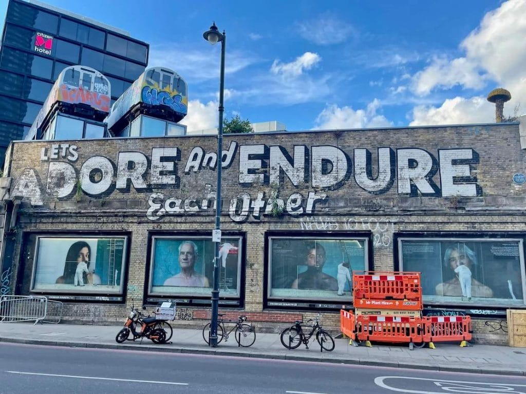 A pastel colour building with four windows with random faces and at the top of the building on the left corner, there are two abandoned train carriages covered in graffiti by Sime and Wendy perched atop two containers. On the right corner, there’s a Cristhiaan Nagel oversized yellow mushroom.