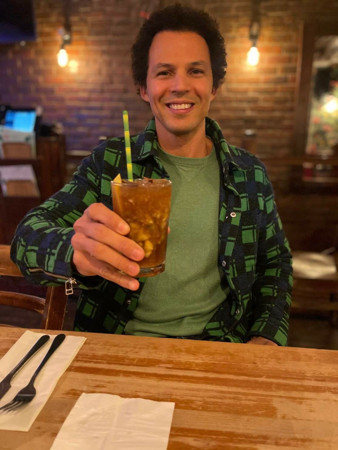 Péricles Rosa usando um suéter verde e uma camisa verde bebendo uma caipirinha de canela com maçã no Restaurante Made in Brasil, Londres