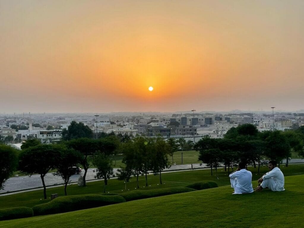 Dos chicos con trajes tradicionales sentados en la hierba y viendo la puesta de sol desde Katara Hills, Doha