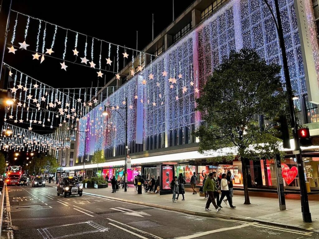 Black cars and red buses running on Oxford Street, which is decorated with Christmas lights and the twinkling John Lewis & Partners building
