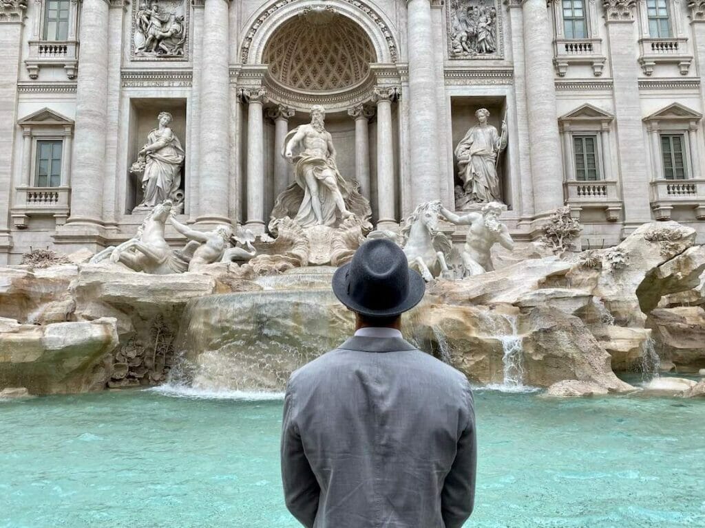 Péricles Rosa wearing a grey heat and a grey suit facing the Fontana de Trevi in Rome, Italy