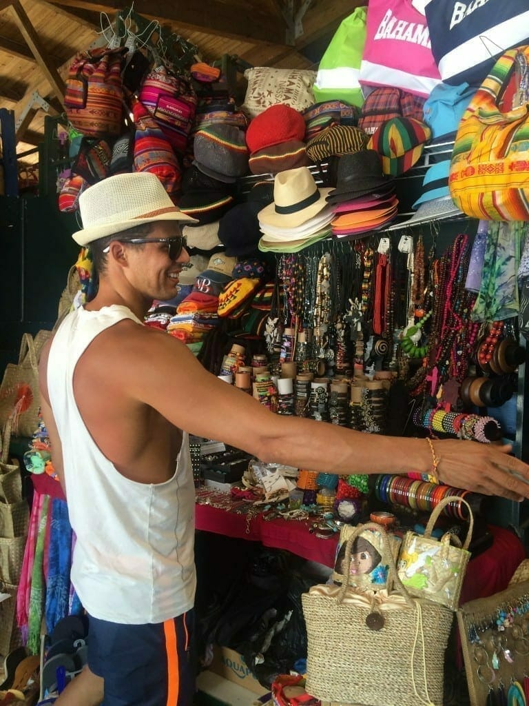 A man wearing a beige hat, sunglasses, a white top and blue shorts shopping at Straw Market, Nassau, Bahamas