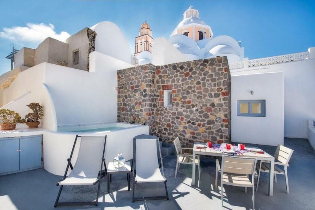 Un apartamento de un dormitorio con bañera de hidromasaje, sillas y una mesa con una iglesia de estilo cicládico como telón de fondo en Anatolia Hotel and Spa, Fira, Santorini  