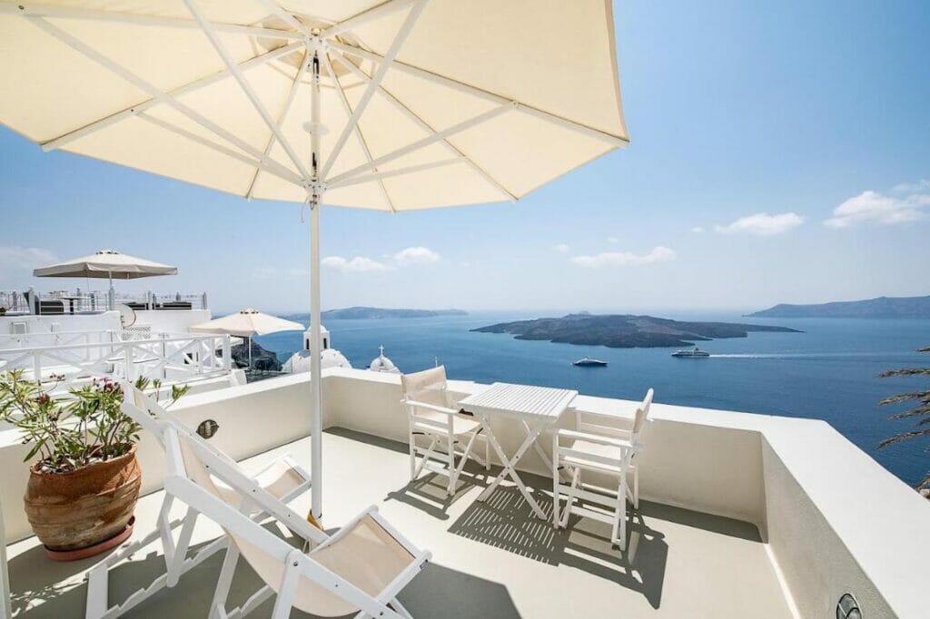 A veranda of a suite with white chairs, a table and an umbrella with views to the caldera and Aegean Sea at Pantelia Suites, Fira, Santorini