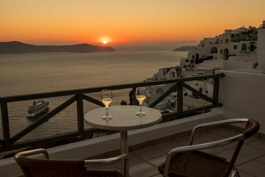 Un balcón con dos sillas, una mesa y dos copas de vino blanco con la puesta de sol como telón de fondo en el mar Egeo, The Last Sunbeam Hotel, Fira, Santorini