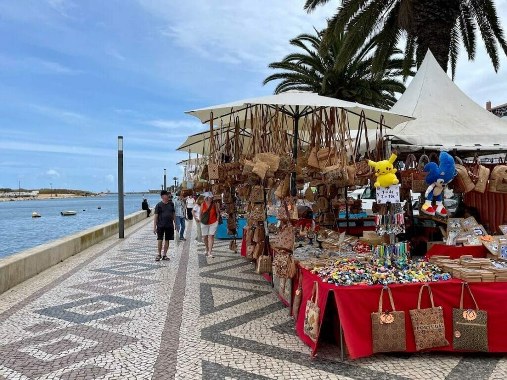 Some people walking on Avenida dos Descobrimentos sidewalk that has lined palm trees and street vendors commercialising local products, Lagos, Algarve, Portugal