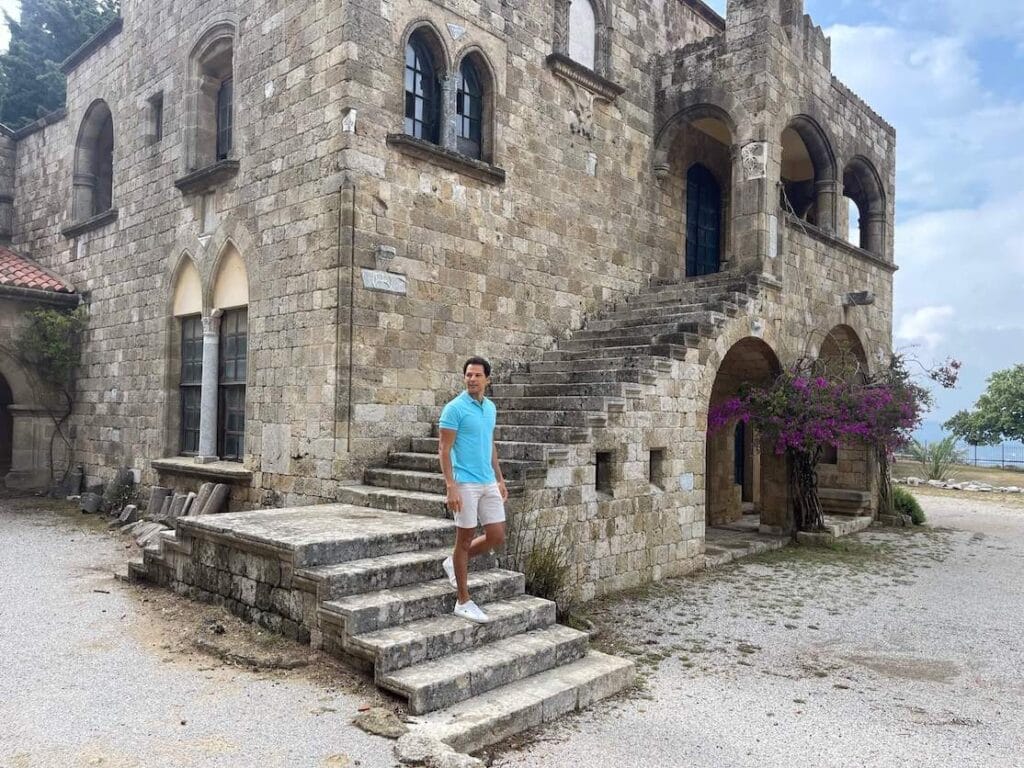 Pericles Rosa vistiendo un polo azul, pantalones cortos beige y zapatillas blancas caminando por las escaleras en el Monasterio de Panagia Filerimos, Rodas, Grecia