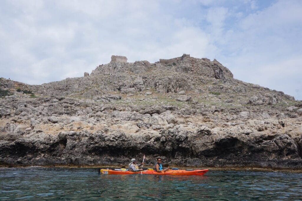 Dos hombres navegando en kayak en el pueblo de Haraki con el Castillo de Feraklos en la cima de una colina al fondo, Rodas, Grecia