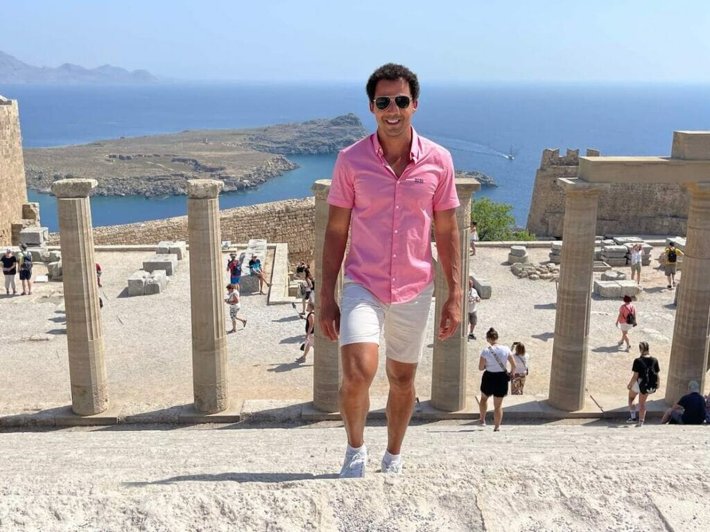 Pericles Rosa wearing sunglasses, salmon shirt, beige shorts and white trainers, posing for a picture ar Lindos Acropolis, Rhodes, Greece