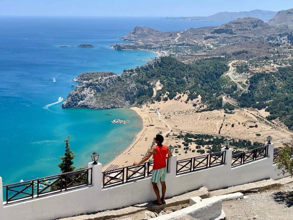 Un hombre vestido con una camiseta sin mangas naranja y pantalones cortos coloridos admirando la vista de la playa de Tsambika desde el monasterio de Tsambika, Rodas, Grecia