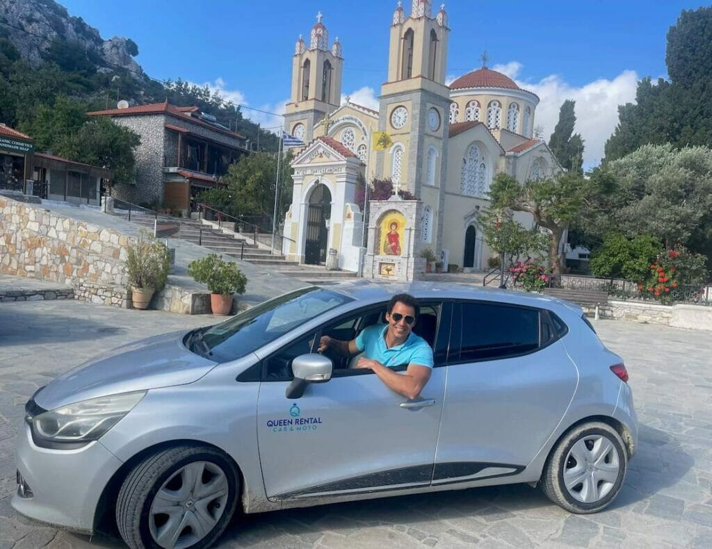 Um homem dentro de um carro alugado com uma igreja ao fundo na aldeia de Siana, Rodes, Grécia