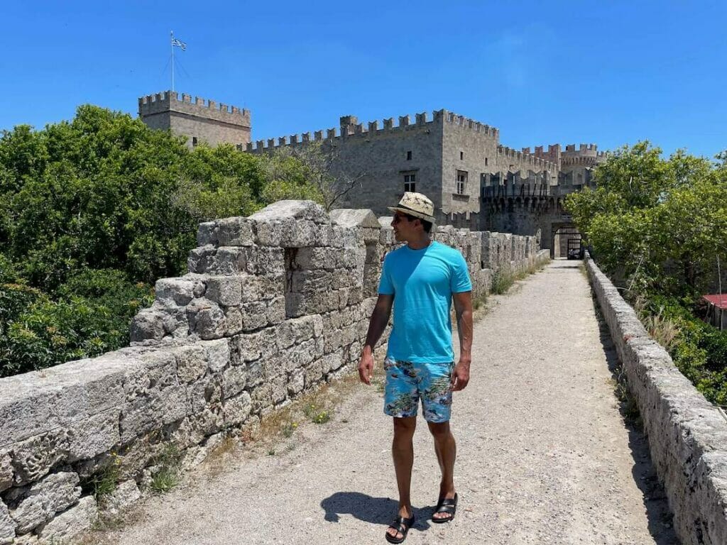 Pericles Rosa wearing a hat, blue T-shirt, colourful shorts and sandals walking on the top of Rhodes city walls, Greece