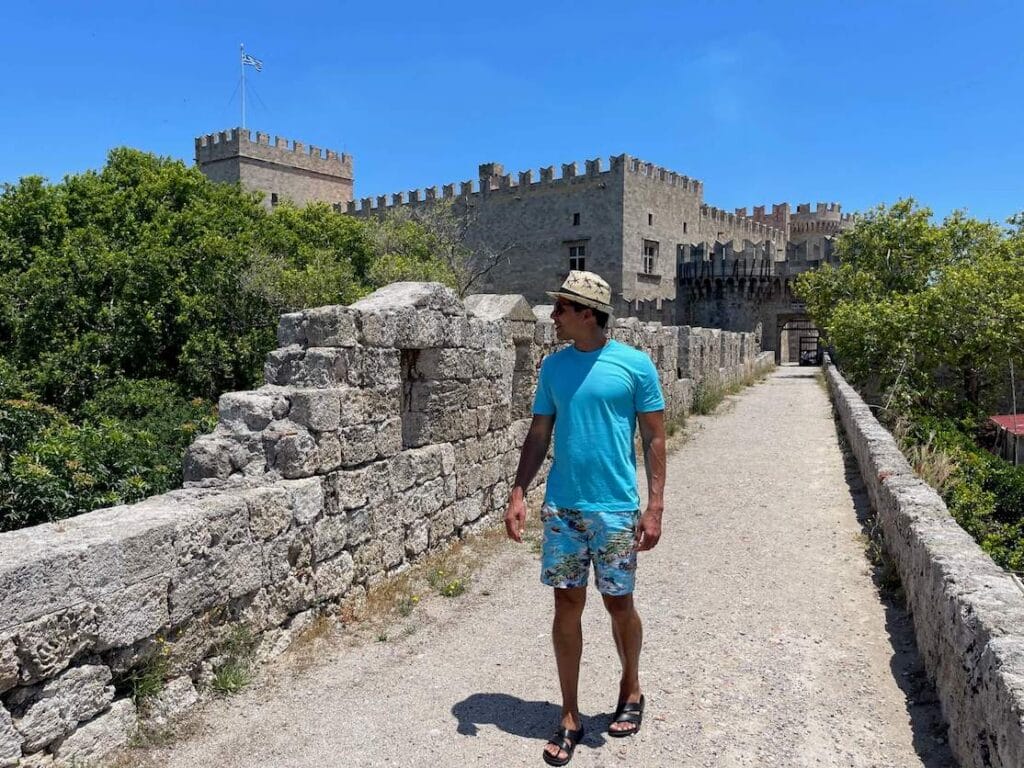 Pericles Rosa con sombrero, camiseta azul, pantalones cortos coloridos y sandalias caminando en la parte superior de las murallas de la ciudad de Rodas, Grecia