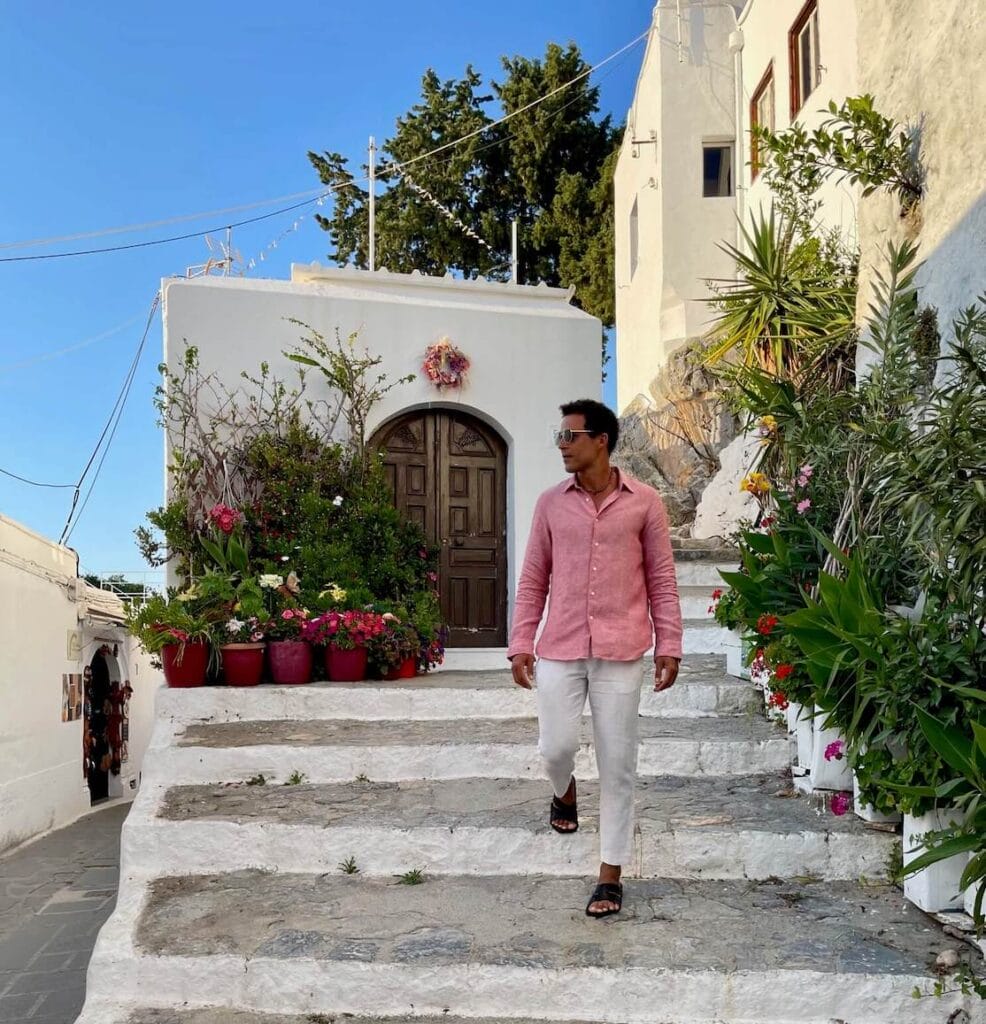 Péricles Rosa vestindo uma camisa salmão de manga comprida, calça bege, óculos escuros e sandálias descendo as escadas em um beco em Lindos, Grécia