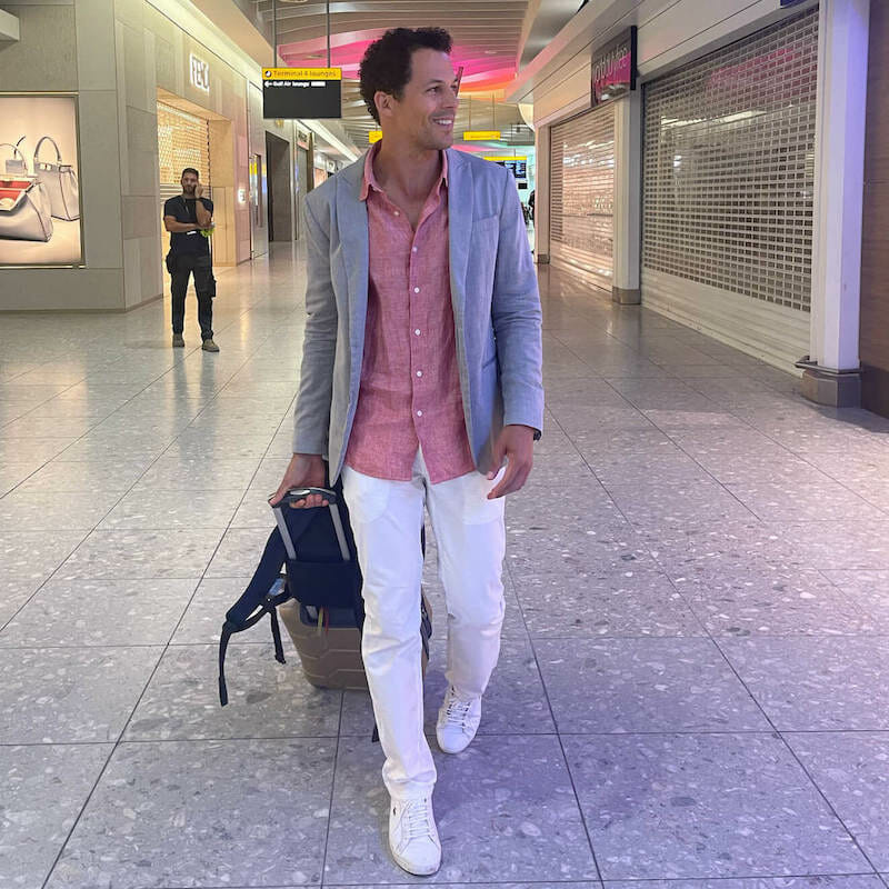 Pericles Rosa wearing a grey blazer, a salmon shirt, white trouser, and a white trainers walking on a terminal of Heathrow Airport carrying a hand luggage. 