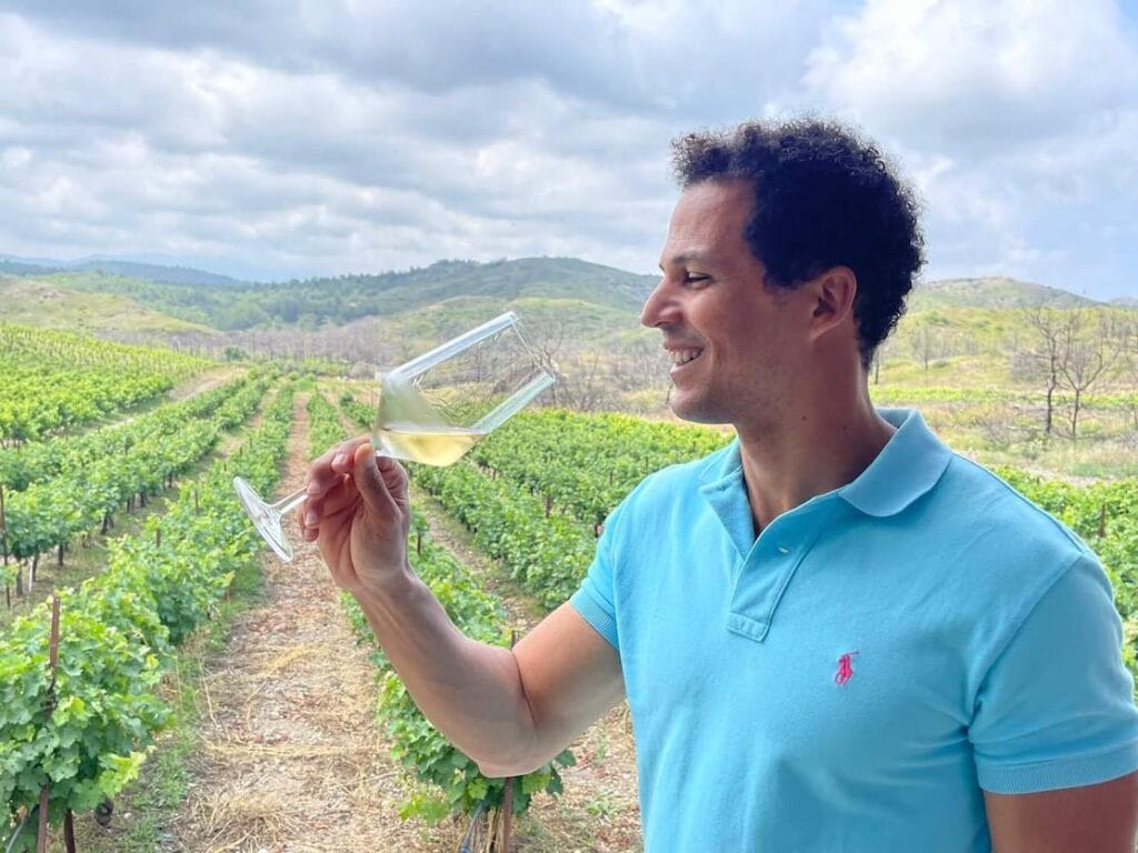 Um homem vestindo um polo azul bebendo um vinho branco na propriedade Anastasia Triantafillou com a vinícola como pano de fundo, Rhodes, Grécia