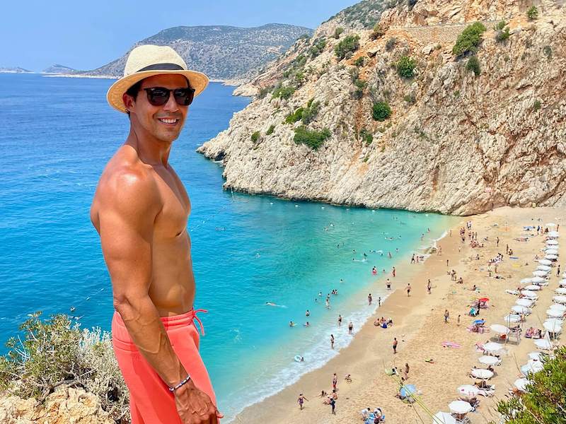 Pericles Rosa wearing sunglasses, a hat and salmon shorts posing for a picture on the of a hill with Kaputas Beach as a backdrop, Kas, Turkey