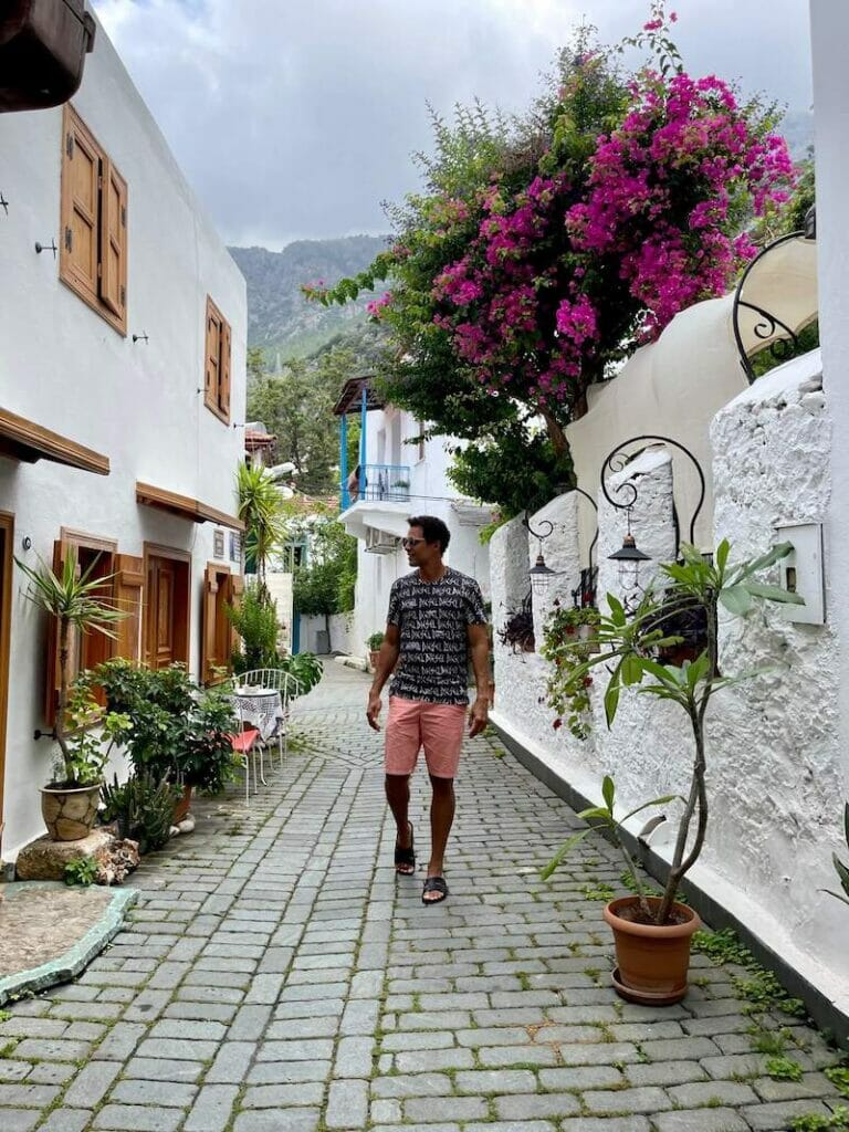 Pericles Rosa wearing sunglasses, a black t-shirt, salmon shorts and black sandals posing for a picture on a whitewashed alley in Kas Old Town, Kas, Turkey