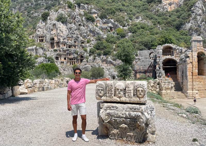 Pericles Rosa wearing sunglasses, a pink t-shirt. beige shorts and white sneakers posing for a picture at the ancient city of Myra, Turkey