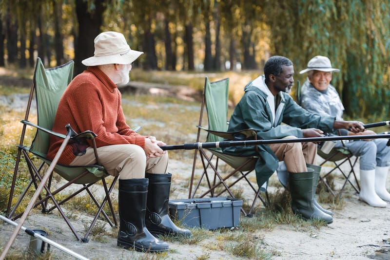 Senior man with fishing rod sitting near toolbox and interracial friends outdoors