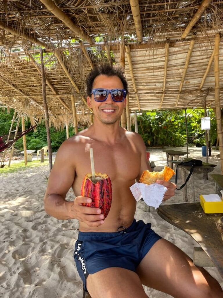 Pericles Rosa wearing blue sunglasses and blue shorts sitting down at Barraca Paraiso on Castelhanos Beach holding a drink on a cacao shell  whit a hand and a pastel with the other hand, Boipeba Island, Bahia, Brazil