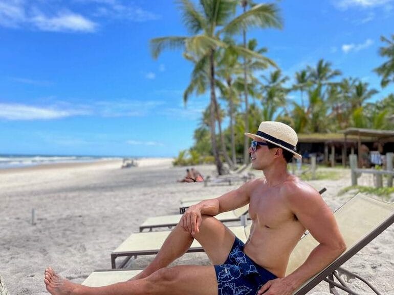 Péricles Rosa usando chapéu bege, óculos escuros e bermuda azul sentada em uma espreguiçadeira com a praia ao fundo no Itacarezinho Restaurante e Lounge, Itacaré, Bahia, Brasil