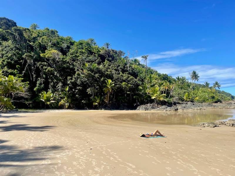 Uma mulher tomando sol na Praia da Camboinha com um morro coberto de floresta ao fundo, Itacaré, Brasil