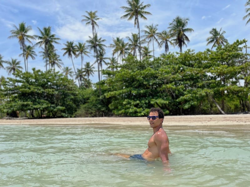 Péricles Rosa usando óculos escuros e short azul na Praia de Bainema, Ilha de Boipeba, Bahia, Brasil