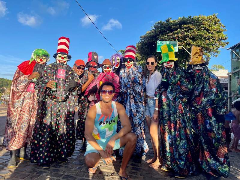 Pessoas usando fantasias de carnaval e três turistas posando para uma foto em Itacaré, Bahia, Brasil