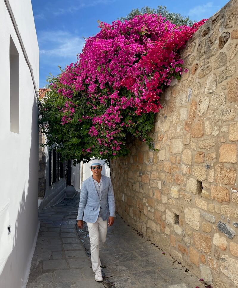 Pericles Rosa con sombrero blanco, blazer azul claro, gafas de sol, pantalones blancos y zapatillas blancas caminando por un callejón con buganvillas en Lindos, Rodas, Grecia