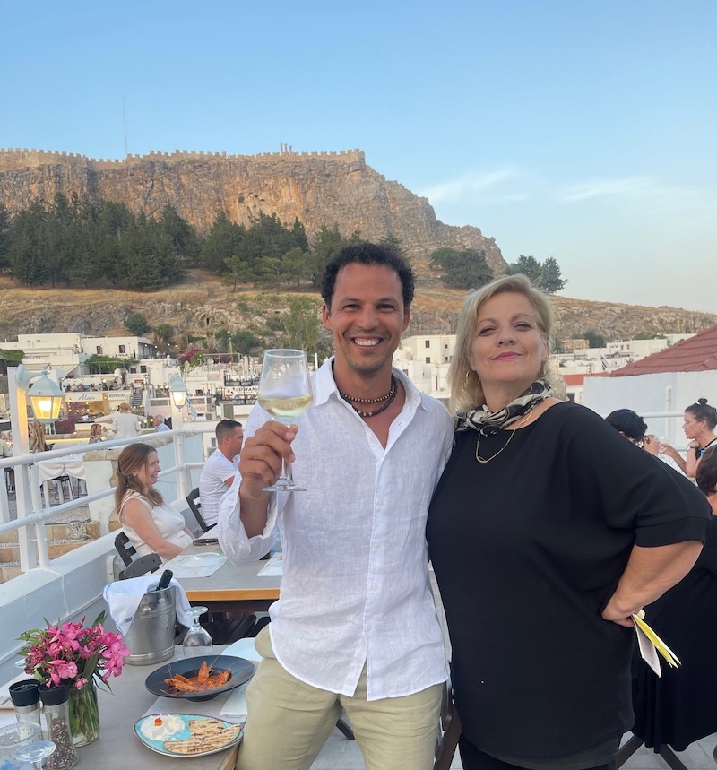 Um homem vestindo uma camisa branca segurando uma taça de vinho branco e uma mulher vestindo preto posando para uma foto no restaurante Kalypso com a Acrópole de Lindos ao fundo, Lindos, Rodes, Grécia