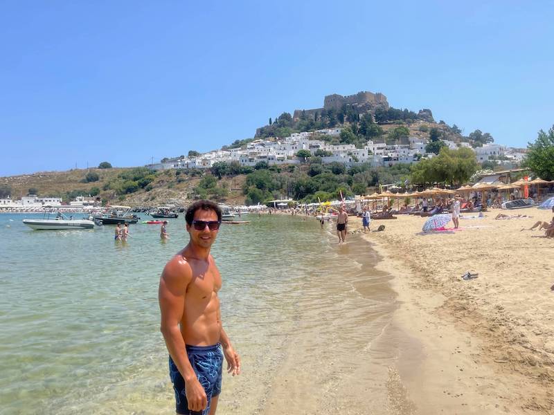 Pericles Rosa posing for a pictures at Lindos Beach with Lindos Town and its Acropolis in the background, Lindos, Rhodes, Greece